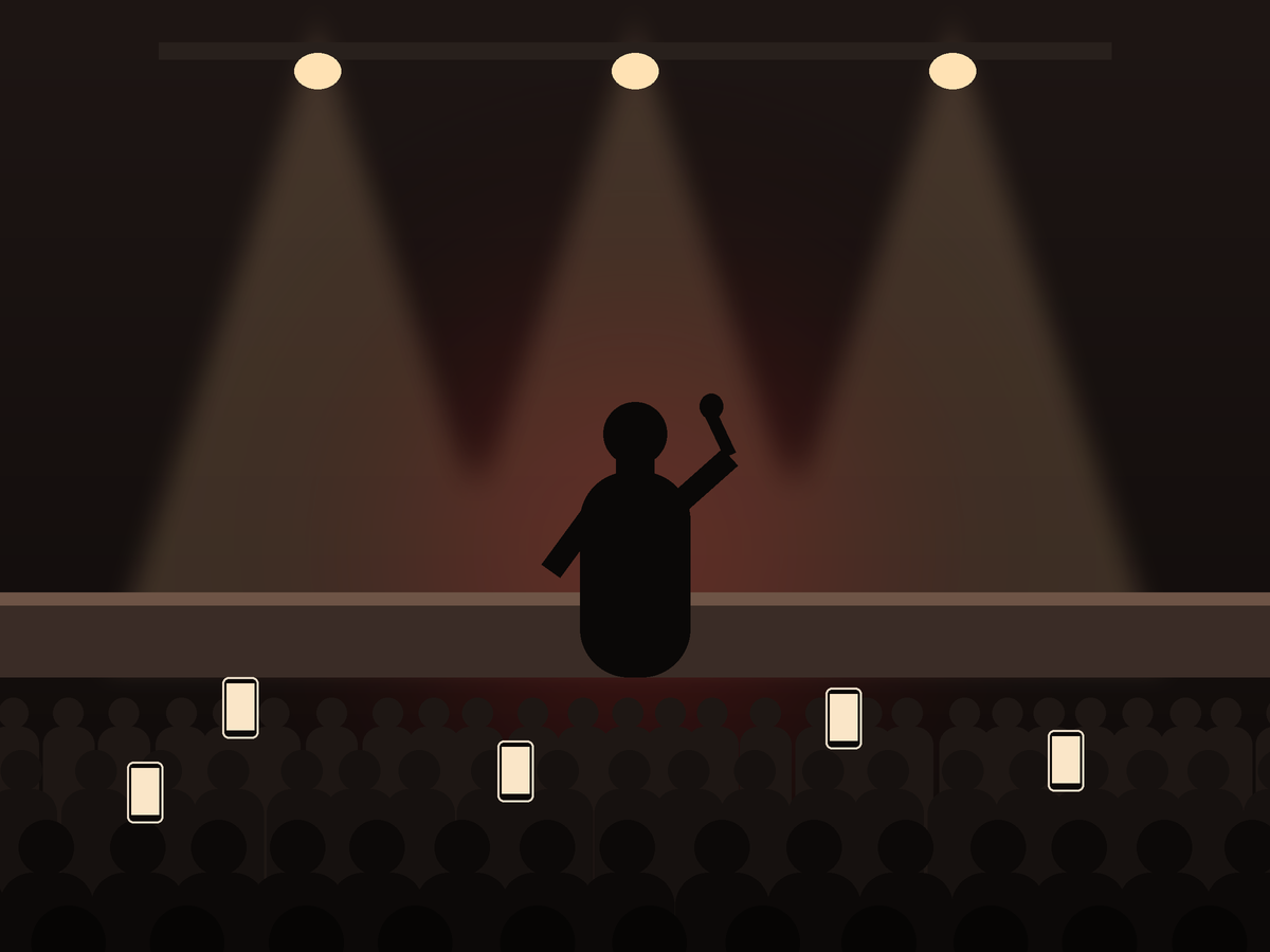A performer on stage under spotlights with an audience filming on their phones